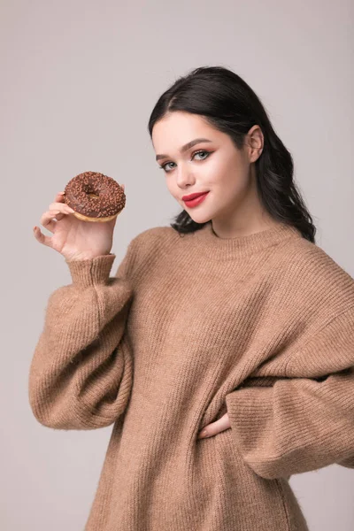 Kırmızı dudaklar çörek ile poz güzel kızla. Uzun saçlı, parlak kırmızı dudaklar, holding lezzetli çörek el ile çok seksi esmer kız kameraya bakıyor. Koyu gri arka plan, kopya alanı