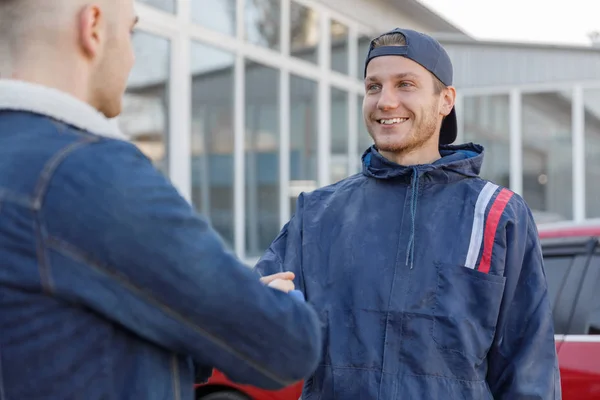 Car service, repair, deal and people concept - mechanic and customer or car owner shaking hands in a workshop. Closeup of an automaster.