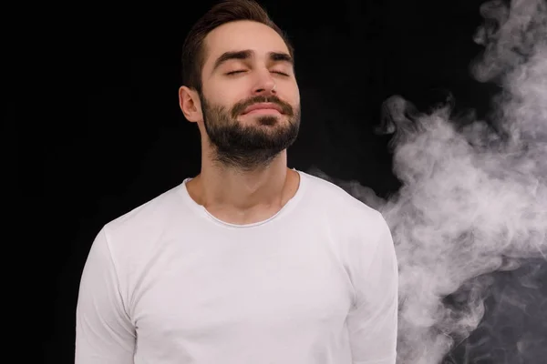 Chico en camiseta blanca inhala humo sobre un fondo negro —  Fotos de Stock