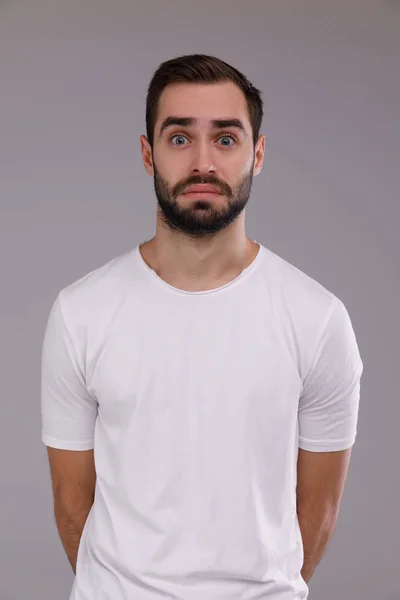 Um homem com uma barba em uma camiseta branca em um fundo cinza surpreso — Fotografia de Stock