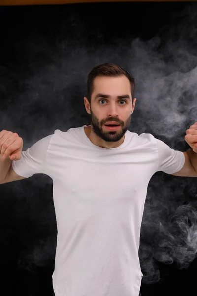 Homem de t-shirt branca sobre fundo preto liberando uma nuvem de fumaça — Fotografia de Stock