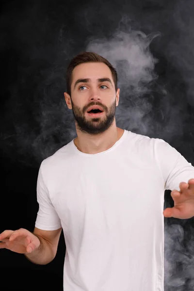 Um jovem em uma camiseta branca em um fundo preto com fumaça olha para o lado — Fotografia de Stock