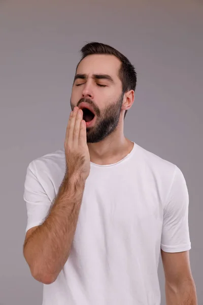 Retrato de um homem em uma camiseta branca que está bocejando contra um fundo cinza . — Fotografia de Stock