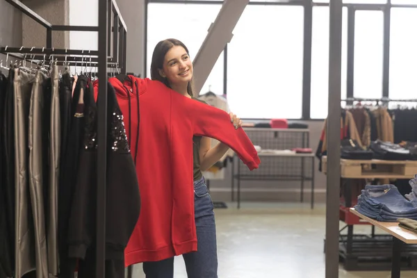 The girl in the store chooses her new things, a red sweatshirt. — Stock Photo, Image