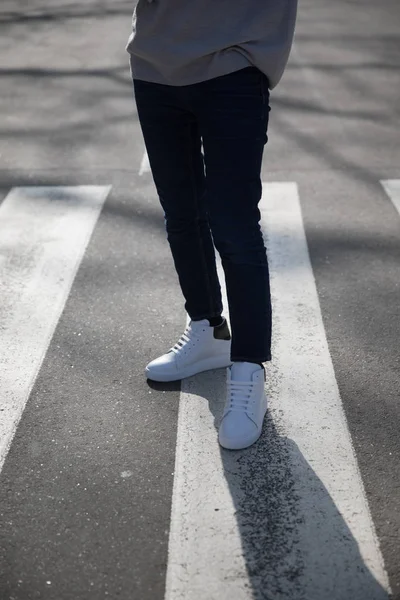 Молодой человек в темных брюках и кроссовках переходит дорогу — стоковое фото