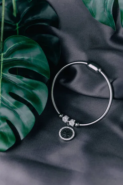 A close-up of a silver bracelet lies on a black background, next to flower sheets, on the bracelet there is a pendant in which is a tree. The concept of jewels.