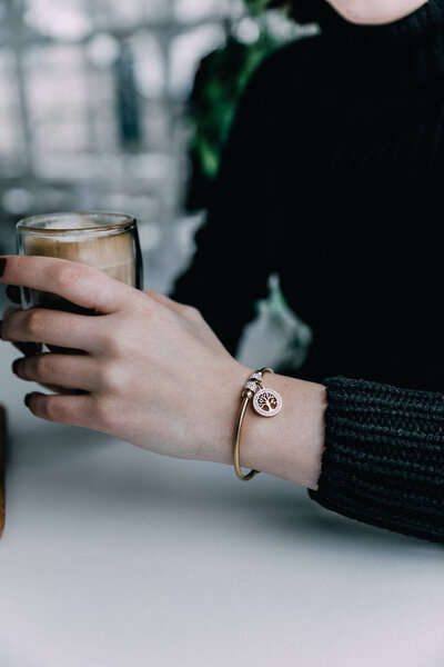 Крупный план золотого браслета на запястье девушки, которая носит черный свитер и держит кофе в руке, на ее браслете есть кулон круглой формы
.