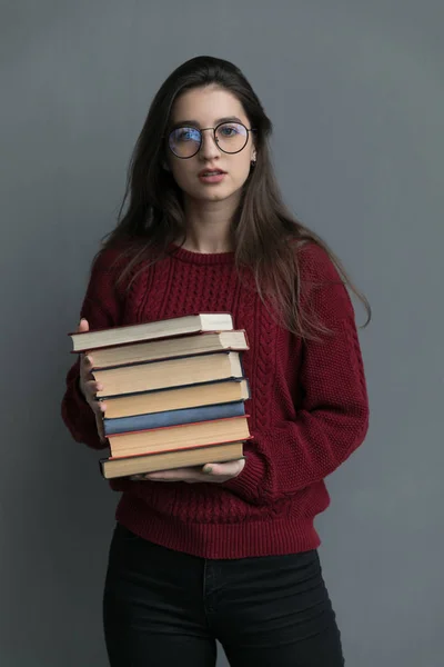 彼女の手に本を保持灰色の背景に ポーズを流れる髪を持つ少女のクローズアップ バーガンディのセーターとメガネを着て ストック画像