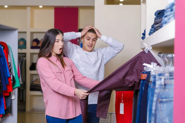 La pareja joven considera la ropa en la tienda. Joven pareja agradable en la tienda con compras —  Fotos de Stock