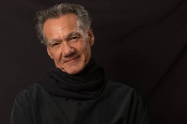 Siyah bir arka planda siyah gömlek ve eşarp takmış gülümseyen yaşlı bir adamın portresi.