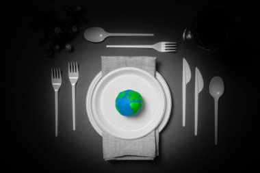 Renkli Dünya gezegeni, siyah beyaz plastik tabaklar, kaşıklar, çatallar ve petrol yağlı şarap kadehleriyle servis masası. Dünya Saati, Dünya Günü, Okyanus Günü ve diğer ECO tarihleri için ekoloji kavramı.