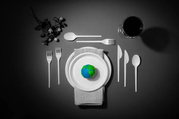 Renkli Dünya gezegeni, siyah beyaz plastik tabaklar, kaşıklar, çatallar ve petrol yağlı şarap kadehleriyle servis masası. Dünya Saati, Dünya Günü, Okyanus Günü ve diğer ECO tarihleri için ekoloji kavramı.
