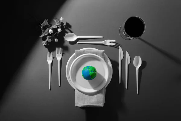 Renkli Dünya gezegeni, siyah beyaz plastik tabaklar, kaşıklar, çatallar ve petrol yağlı şarap kadehleriyle servis masası. Dünya Saati, Dünya Günü, Okyanus Günü ve diğer ECO tarihleri için ekoloji kavramı.