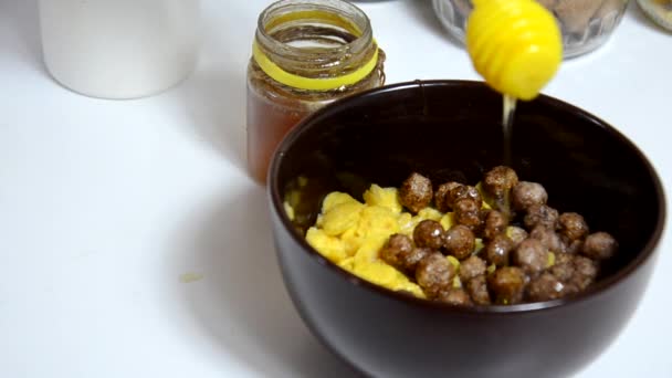 Honig zum Frühstückszerealien geben, Schokokugeln in eine braune Schüssel geben. gesundes Frühstück. Diätschale und Löffel mit Cornflakes isoliert auf weißem Hintergrund — Stockvideo