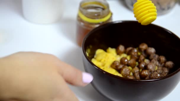 把蜂蜜倒进早餐麦片里，巧克力球放在棕色的碗里。健康的早餐。饮食碗和勺子与玉米片隔离在白色背景 — 图库视频影像