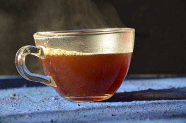 Bir pencerenin önünde koyu mavi yüzeye yerleştirilen şeffaf kahve fincanı. buhar ile karanlık bir arka plan üzerinde sıcak siyah kahve ile cam fincan