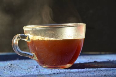 Bir pencerenin önünde koyu mavi yüzeye yerleştirilen şeffaf kahve fincanı. buhar ile karanlık bir arka plan üzerinde sıcak siyah kahve ile cam fincan