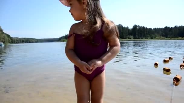 Fetița drăguță intră în apă pentru a înota în nisipul de pe plajă. într-un costum de baie violet. Pe un lac sau pe mare. Vară, vacanţă, vacanţă, copilărie fericită. Fata frumoasă în picioare în apă . — Videoclip de stoc