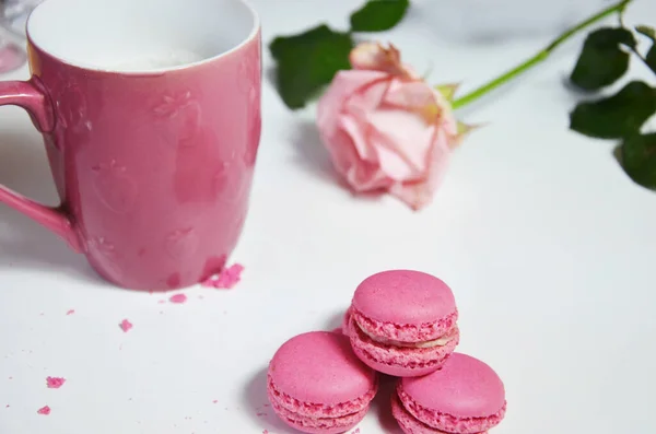 Drei rosa Mandelmakronen und ein Becher mit Kaffee auf weißem Holzgrund neben einer blassrosa Rose. Guten Morgen Konzept — Stockfoto