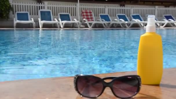 Okulary przeciwsłoneczne i filtry przeciwsłoneczne wokół basenu na tle niebieskiej wody. Ciemne przyciemnione okulary przeciwsłoneczne z kroplami wody odbijającej basen. Jasny słoneczny dzień. — Wideo stockowe