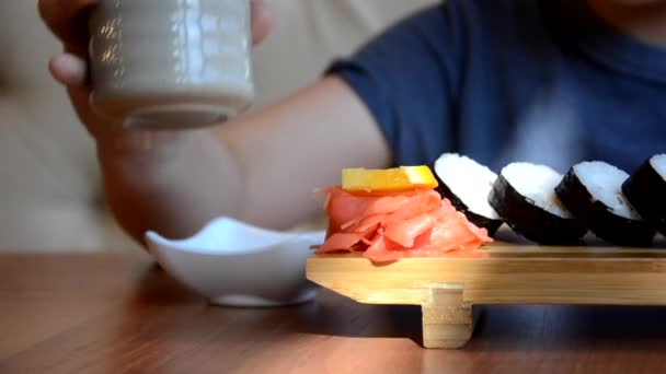 Ένας έφηβος παίρνει ρολό σούσι με κινέζικα μπαστούνια μπαμπού και το ρίχνει στη σάλτσα σόγιας, παίρνει ένα κομμάτι ροζ τζίντζερ. Ιαπωνική κουζίνα σε ένα λευκό πιάτο πορσελάνης δίπλα στο πράσινο wasabi τζίντζερ και σάλτσα — Αρχείο Βίντεο