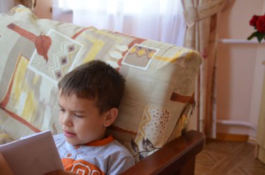 Çocuk evde turuncu tişörtlü bir koltukta oturan bir kitap okuyor. ders çalış. Hobi. Macera. Genç adam kitap okuyor.