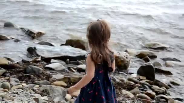 Маленька дівчинка в блакитному вечірньому одязі і рожевий капелюх прогулюється уздовж моря на кам'яному березі. прогулянки, відпустка, подорожі, сім'я — стокове відео
