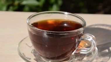 Yeşil yaprakların arka planında şeffaf bardakta çay. Günaydın, açık hava kafe, enerji takviyesi. Çayın içine çay dökülüyor.