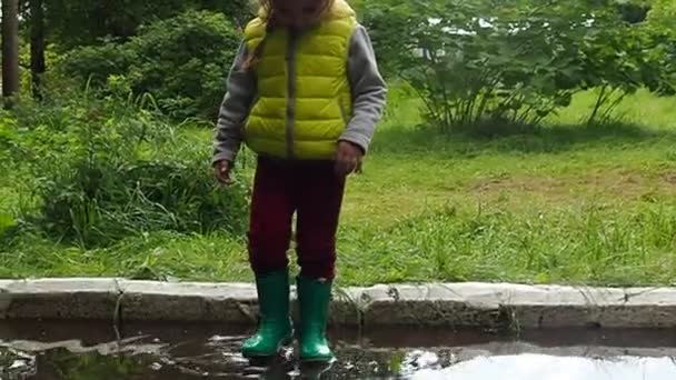 Petite fille mignonne drôle marche sous la pluie avec un parapluie rouge, en bottes en caoutchouc vert à travers les flaques d'eau. Rire, s'amuser. fille portant un manteau imperméable jaune jouant sur une chaude journée d'automne ou d'été — Video