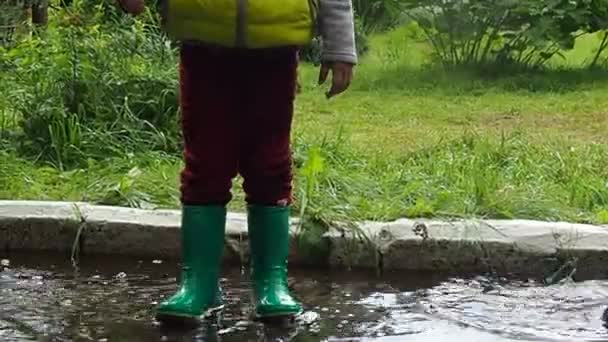 Petite fille mignonne drôle marche sous la pluie avec un parapluie rouge, en bottes en caoutchouc vert à travers les flaques d'eau. Rire, s'amuser. fille portant un manteau imperméable jaune jouant sur une chaude journée d'automne ou d'été — Video