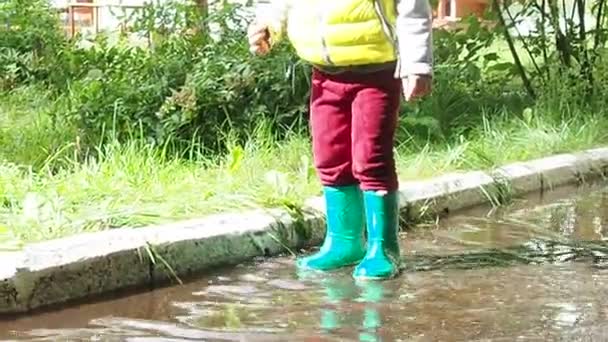 Malá roztomilá holčička chodí v dešti s červeným deštníkem, v zelených gumových botách mezi loužemi. Směje se, baví se. dívka na sobě žlutý nepromokavý kabát hrát na teplý podzim nebo letní den — Stock video