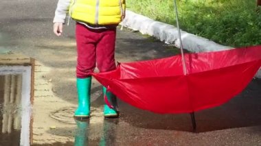 Küçük sevimli kız yağmurda kırmızı şemsiyeli yeşil lastik botlarıyla su birikintilerinde yürüyor. Gülün, eğlenin. Sıcak bir sonbahar ya da yaz gününde oynayan sarı su geçirmez ceket giyen bir kız.