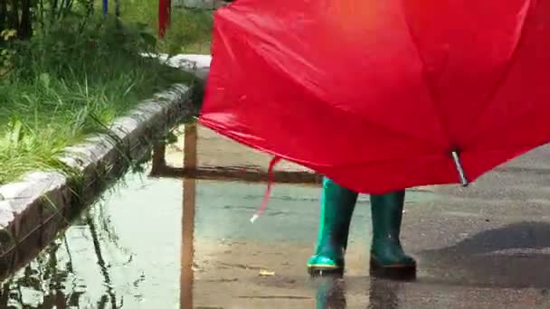 かわいい女の子は赤い傘で雨の中を歩く緑のゴム製のブーツで水たまりを通って。うわ楽しい。暖かい秋や夏の日に黄色の防水コートを着ている女の子 — ストック動画