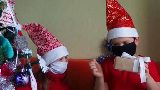 Κοριτσάκι και αγόρι με μάσκα, Χριστουγεννιάτικο στεφάνι και πανδημία έννοια, αδελφός και αδελφή ξεπακετάρει δώρα, έννοια του νέου έτους, οικογένεια — Αρχείο Βίντεο
