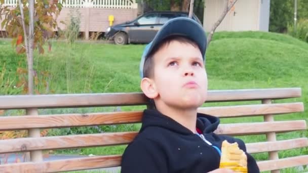 Adorable niño sentado en el banco con lonchera en el parque. Comida saludable y aperitivos para niños. Niño comiendo su desayuno o almuerzo al aire libre después del partido de fútbol — Vídeo de stock