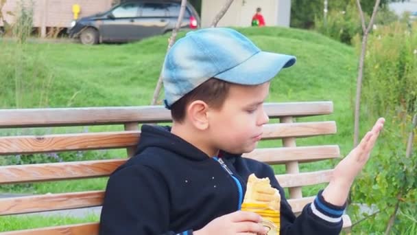 Adorable niño sentado en el banco con lonchera en el parque. Comida saludable y aperitivos para niños. Niño comiendo su desayuno o almuerzo al aire libre después del partido de fútbol — Vídeo de stock