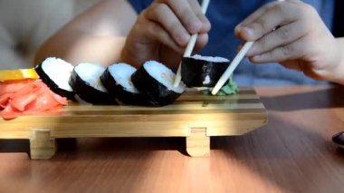 Genç bir çocuk Çin bambu çubuklarıyla suşi rulosunu alıp soya sosunun içine düşürüyor ve bir parça pembe zencefil alıyor. Yeşil wasabi zencefil ve sosun yanında beyaz porselen tabakta Japon mutfağı.