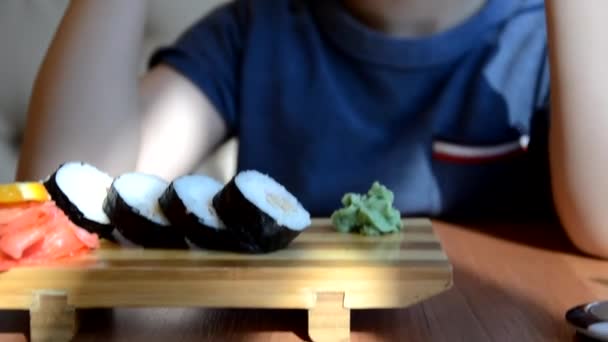 Um adolescente pega o rolo de sushi com paus de bambu chineses e deixa cair no molho de soja, pega um pedaço de gengibre rosa. Cozinha japonesa em um prato de porcelana branca ao lado de gengibre wasabi verde e molho — Vídeo de Stock