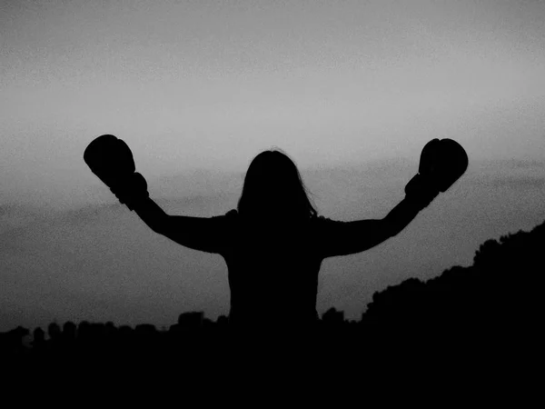 A girl with boxing gloves in the hand. Silhouettes of a boxer woman with a raised hand up. Sunset. Sportswoman holding boxing gloves in the box. Winner