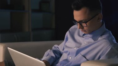 Karanlık bir ofiste bir dizüstü bilgisayar, bir adam oturuyor. Adam geç saatlere kadar çalışıyor