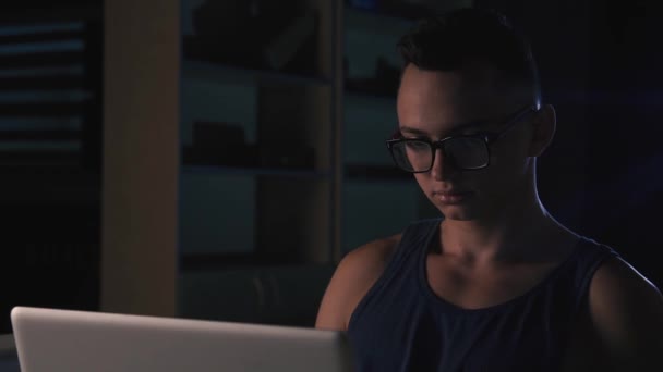 一个戴着眼镜的年轻人在黑暗的房间里在笔记本电脑后面工作 — 图库视频影像