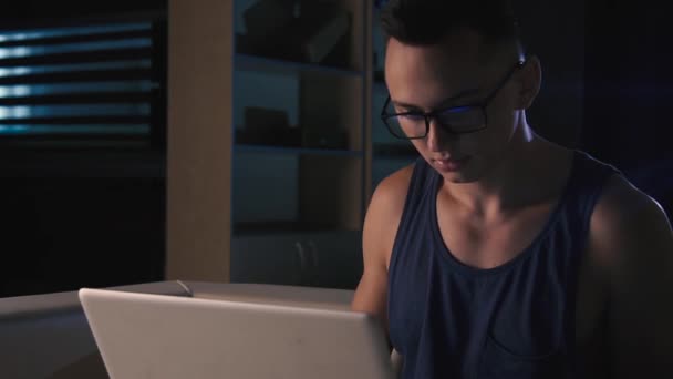 Gözlüklü genç bir adam karanlık bir odada bir dizüstü bilgisayarın arkasında çalışıyor. — Stok video