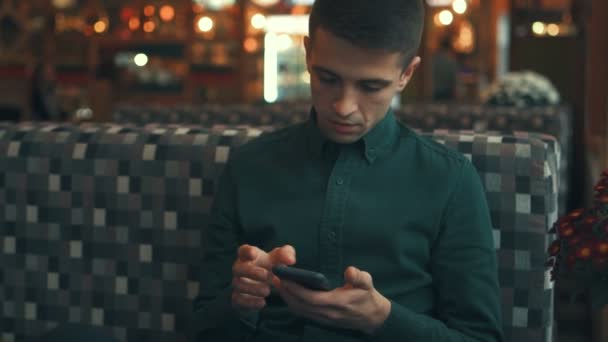 Chico guapo, mirando a su alrededor y utilizando su teléfono inteligente, sentado en un café, charlando. Aspecto elegante, humor juguetón . — Vídeos de Stock