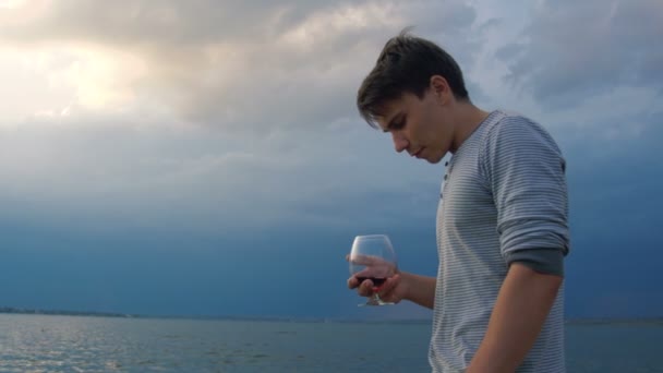 Молодой человек пьет вино на закате у моря — стоковое видео