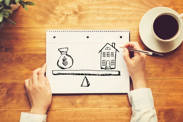 Dům a peníze na stupnici s osobou držící pero — Stock fotografie