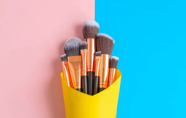 premium makeup brushes on blue and pink background in yellow paper cone, creative cosmetics flat lay, tools for make up, copy space