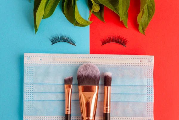 a face made from false eyelashes, a protective face mask and make up brushes on a colored red and blue background, a creative flat lay during a pandemic