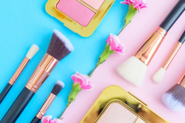 premium makeup brushes, eye shadow palette and flowers on a colored blue and pink background, creative cosmetics flat lay with diagonal composition