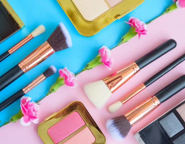 premium makeup brushes, eye shadow palette and flowers on a colored blue and pink background, creative cosmetics flat lay with diagonal composition
