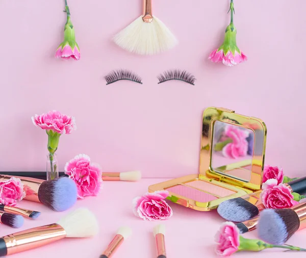 face with false eyelashes and flowers, premium makeup brushes on a colored pink background, creative cosmetics flat lay